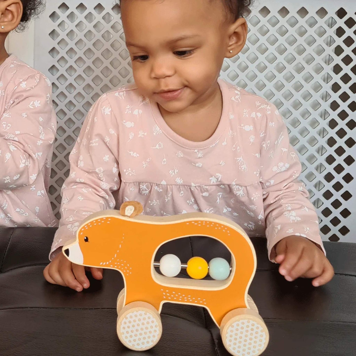 A young child wearing a pink dress engaging with the Push Along Bear, sliding the beads while seated at a black table.
