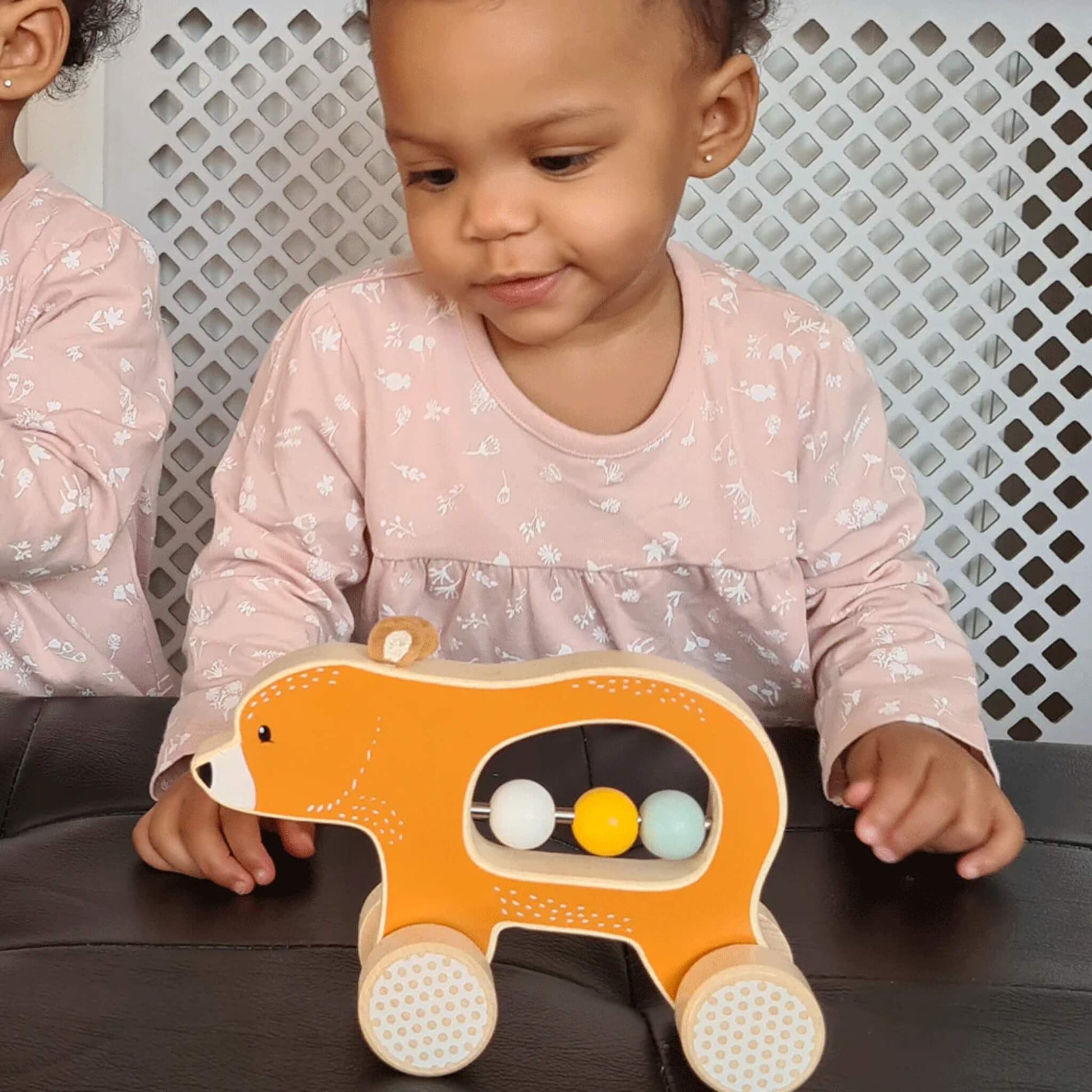 A young child wearing a pink dress engaging with the Push Along Bear, sliding the beads while seated at a black table.