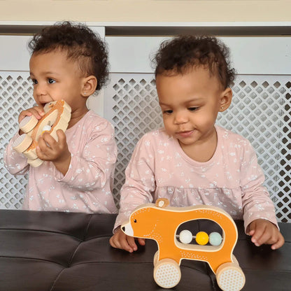 Two toddlers in matching pink outfits, one playing with the Push Along Bear while the other explores another Bigjigs toy, both seated at a table.