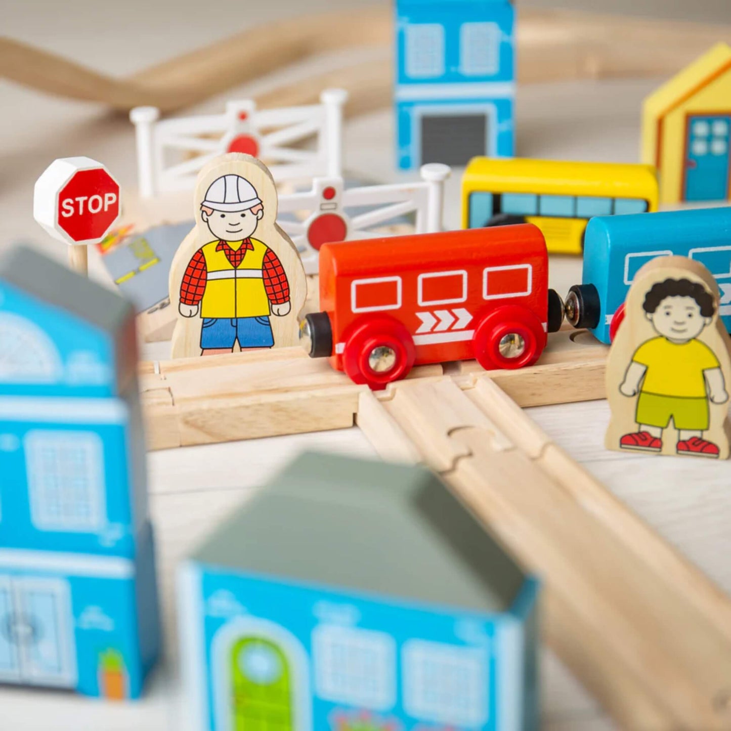 Wooden railway crossing from the Bigjigs Toys Town & Country Train Set, showing a red carriage, construction worker figure, stop sign, and crossing gate surrounded by houses and children figures.