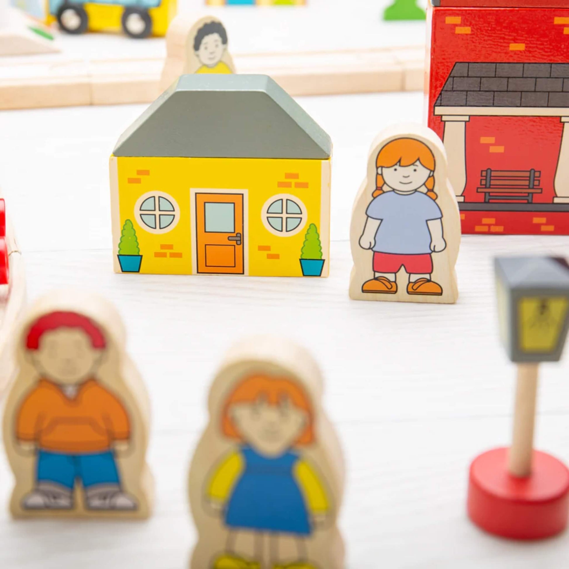 Yellow house and girl play figure surrounded by other wooden characters from the Bigjigs Toys Town & Country Train Set.