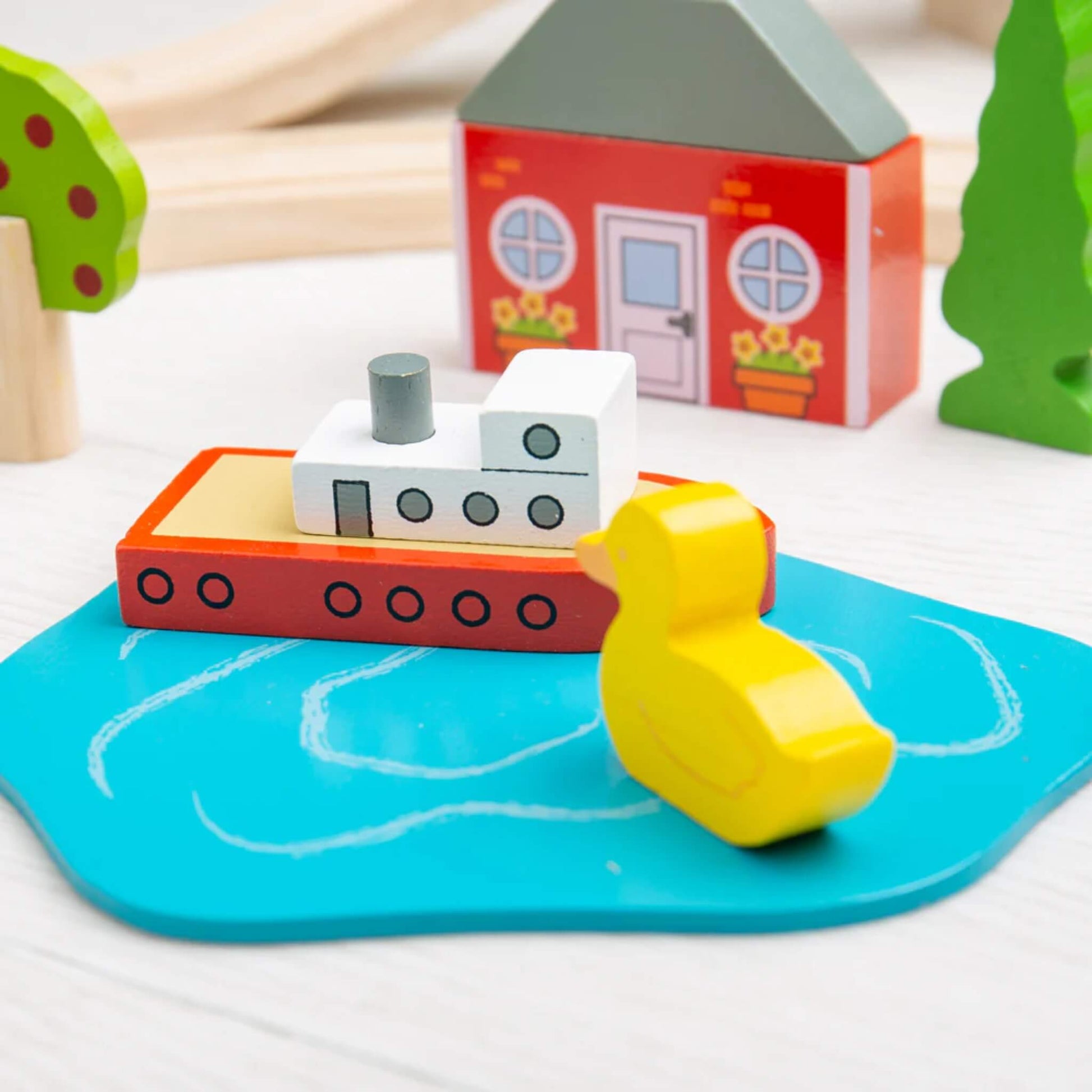Miniature duck and red boat on a wooden pond accessory from the Bigjigs Toys Town & Country Train Set, with background houses and trees.
