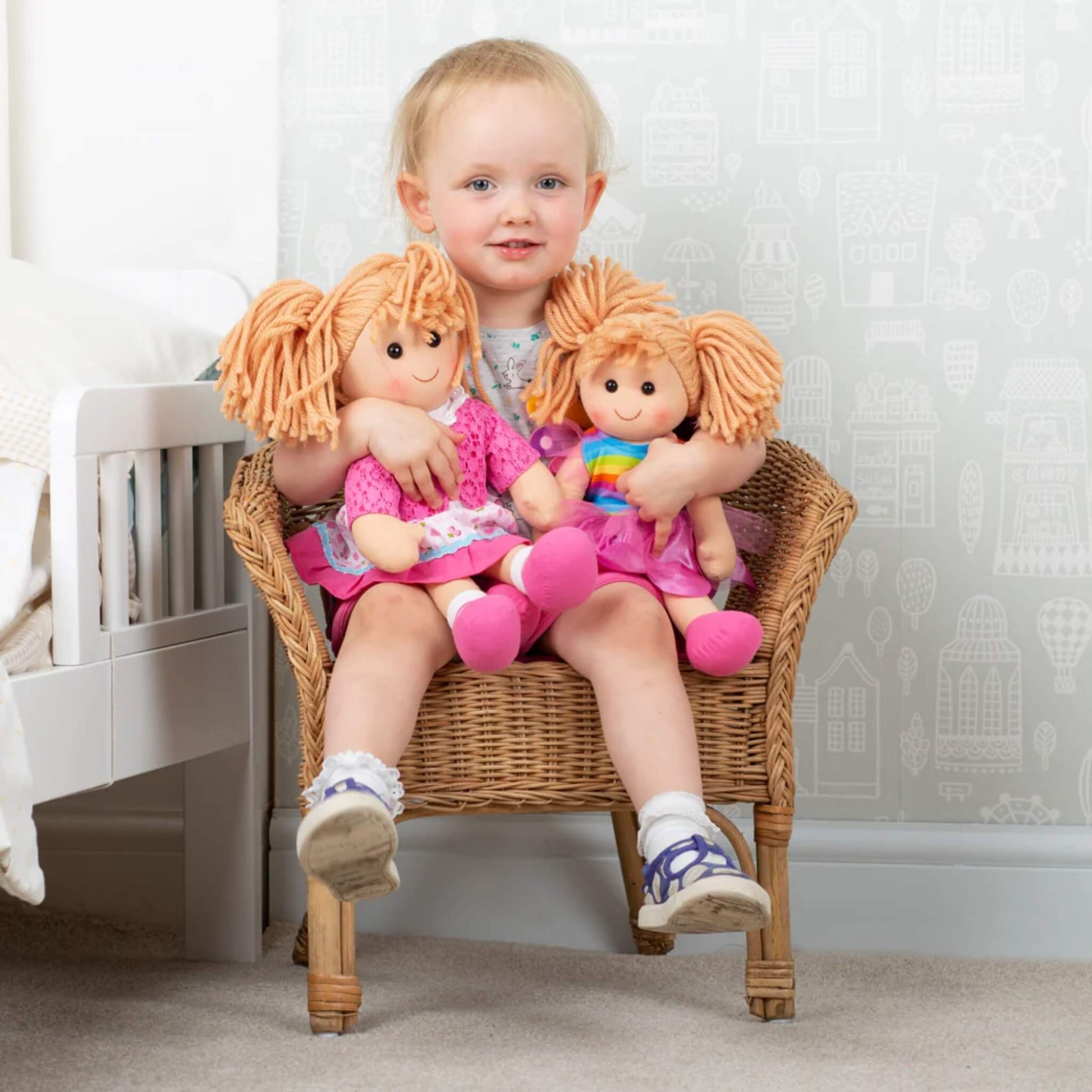 A toddler sitting in a wicker chair cuddling two Bigjigs soft dolls, including Becky in her signature outfit.