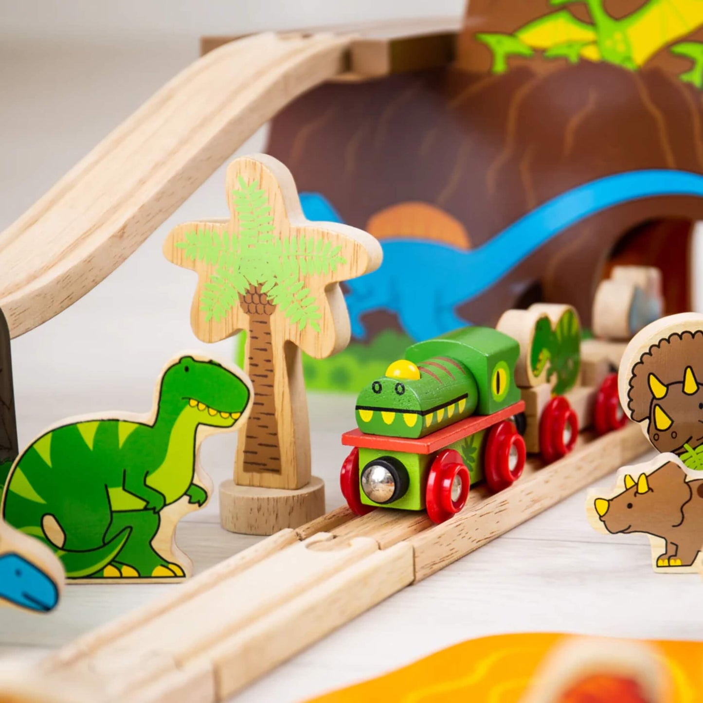 Close-up of the wooden dinosaur train travelling under a raised bridge near palm trees and bright dinosaur figures in a volcano-themed play area.
