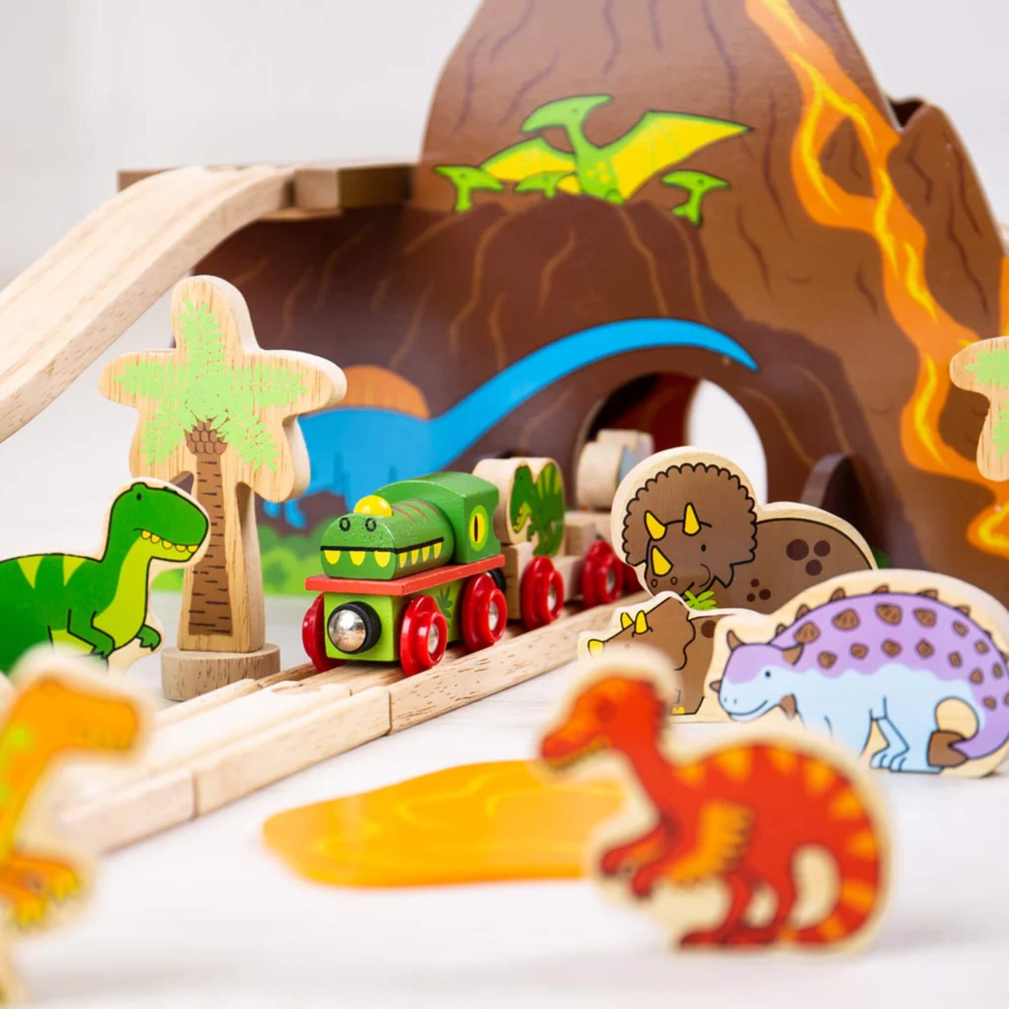 Dinosaur-themed green train emerging from the volcano tunnel, surrounded by wooden dinosaur characters and palm trees in a prehistoric scene.