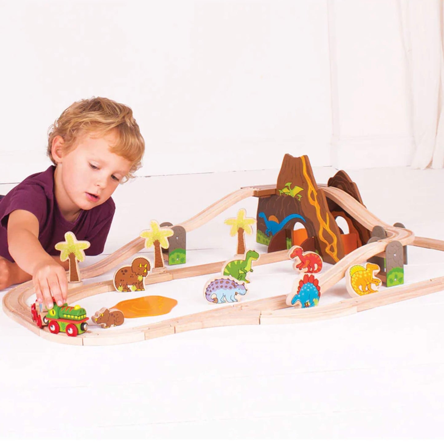 Young child playing with the Bigjigs Toys Dinosaur Railway Set, featuring volcano tunnel, wooden tracks, and dinosaur figures in a bright playroom setting.