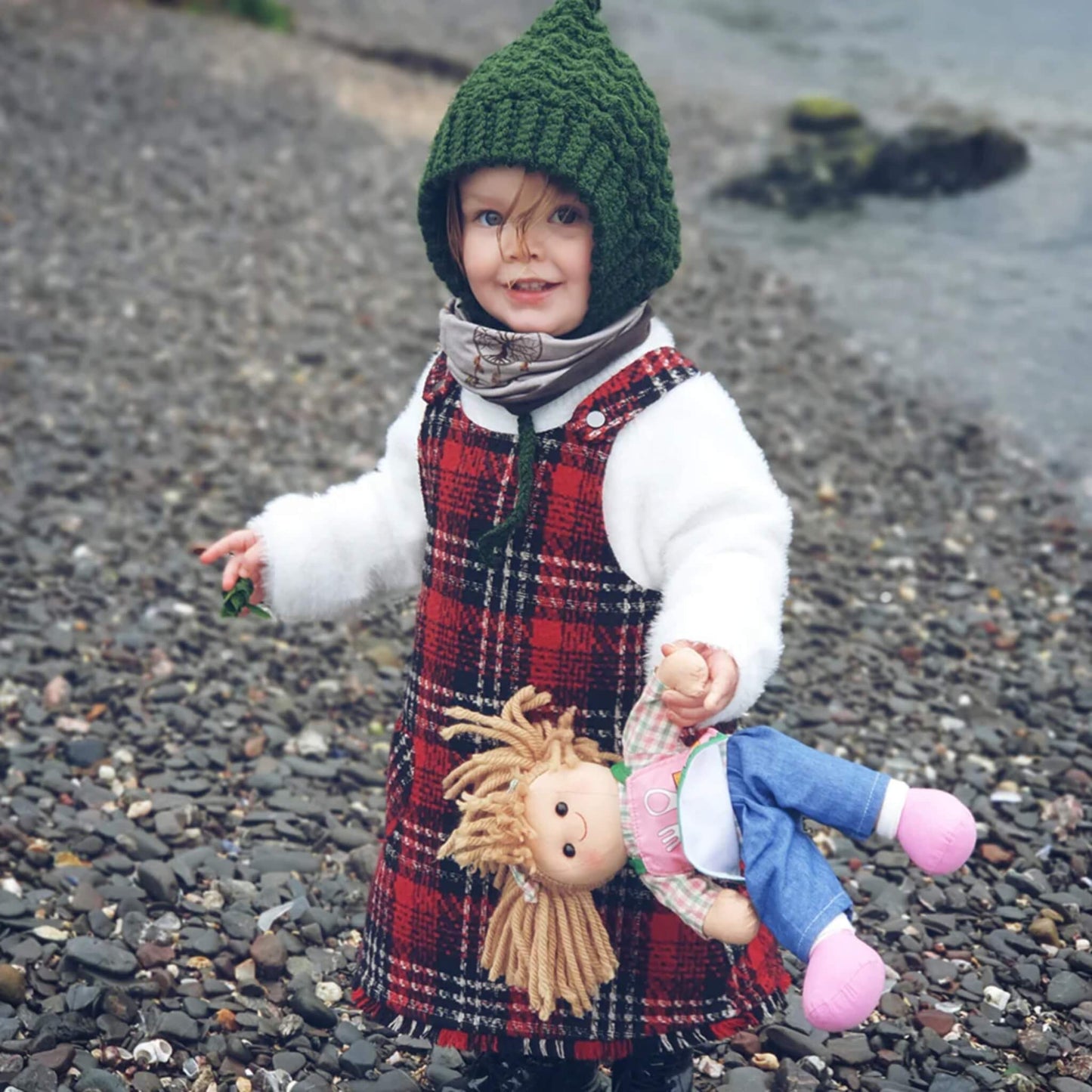 Child in a red check dress and green knit bonnet holding Elsie Doll by the arm while standing on a pebbled beach.