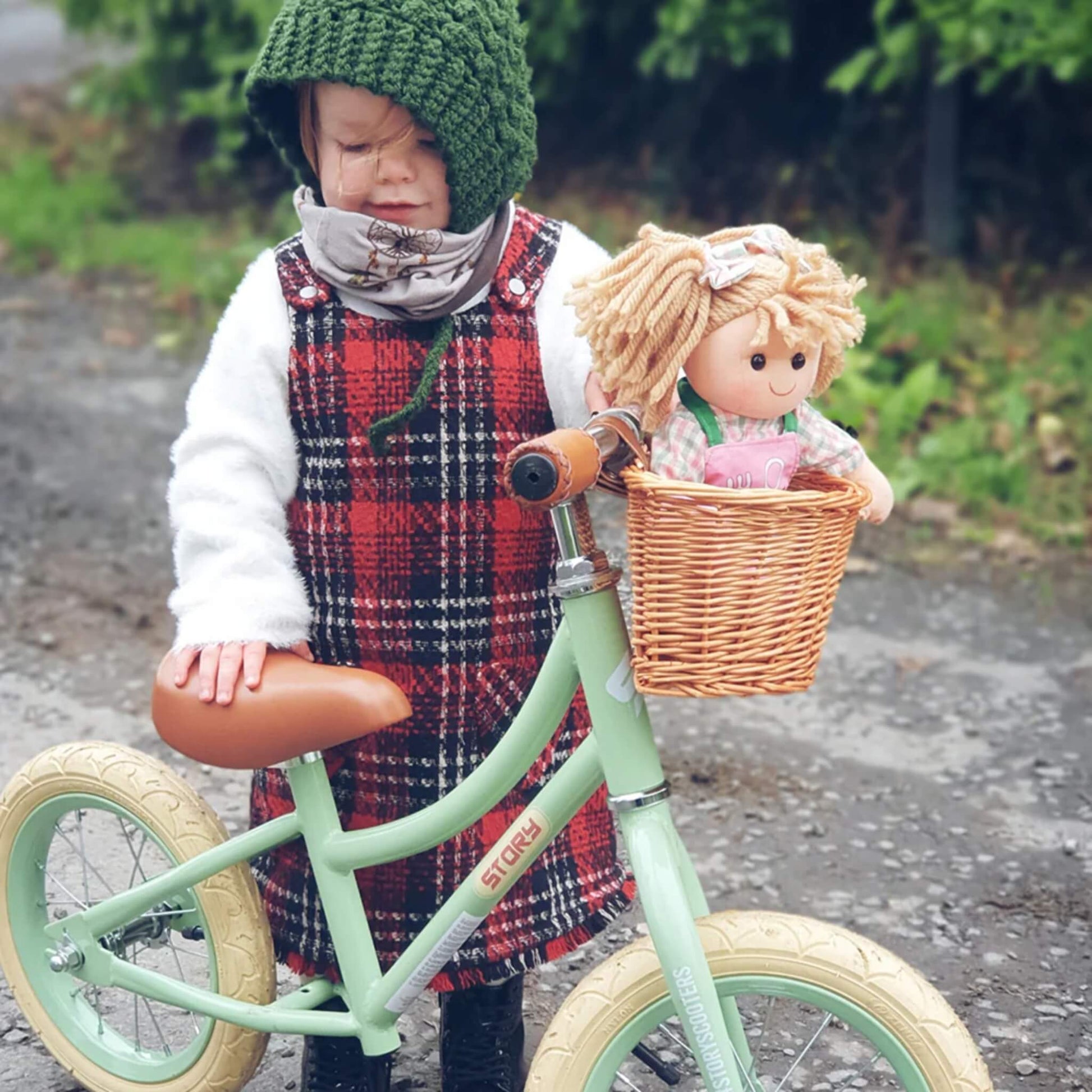 Toddler in a tartan dress and green bonnet standing beside a balance bike, with Elsie Doll sitting in the front wicker basket.