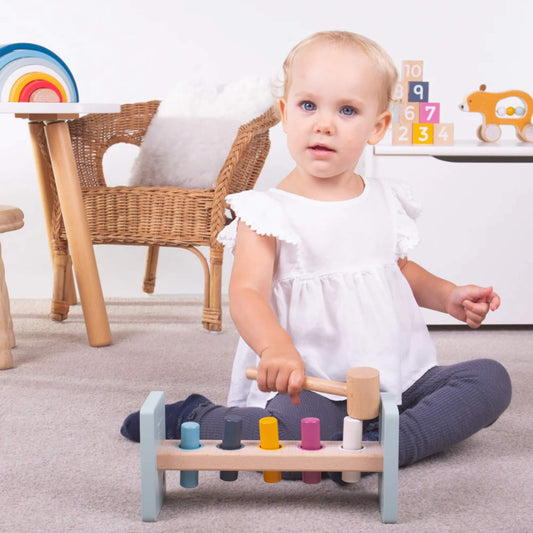 Toddler sitting cross-legged on a carpeted floor, holding the Bigjigs Toys Hammer Bench and preparing to strike a peg with the wooden hammer.