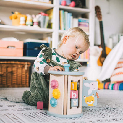 Baby playing on the floor with the Bigjigs Rolling Activity Sorter, exploring the flip book and gear features in a cosy, toy-filled room.