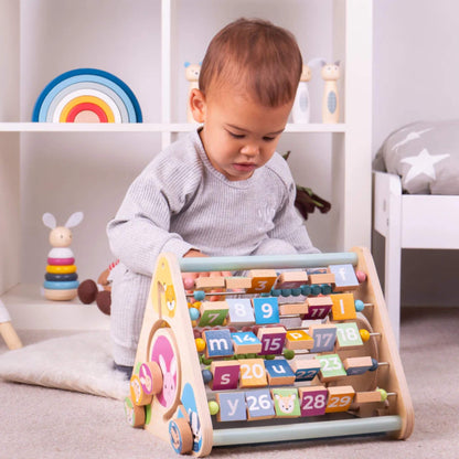 Toddler sitting on a nursery floor, turning blocks on the FSC® wooden activity centre with focused engagement.