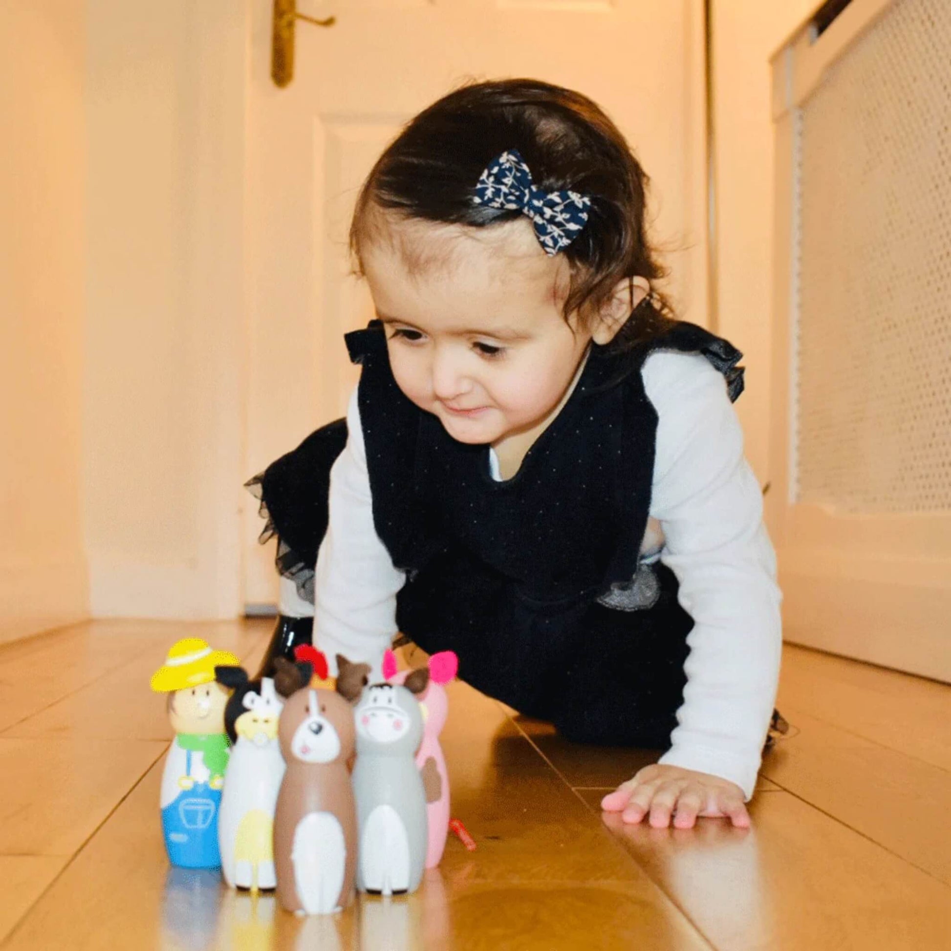 A young child playing indoors, smiling at a line of wooden farm skittles arranged neatly on the floor in front of her.
