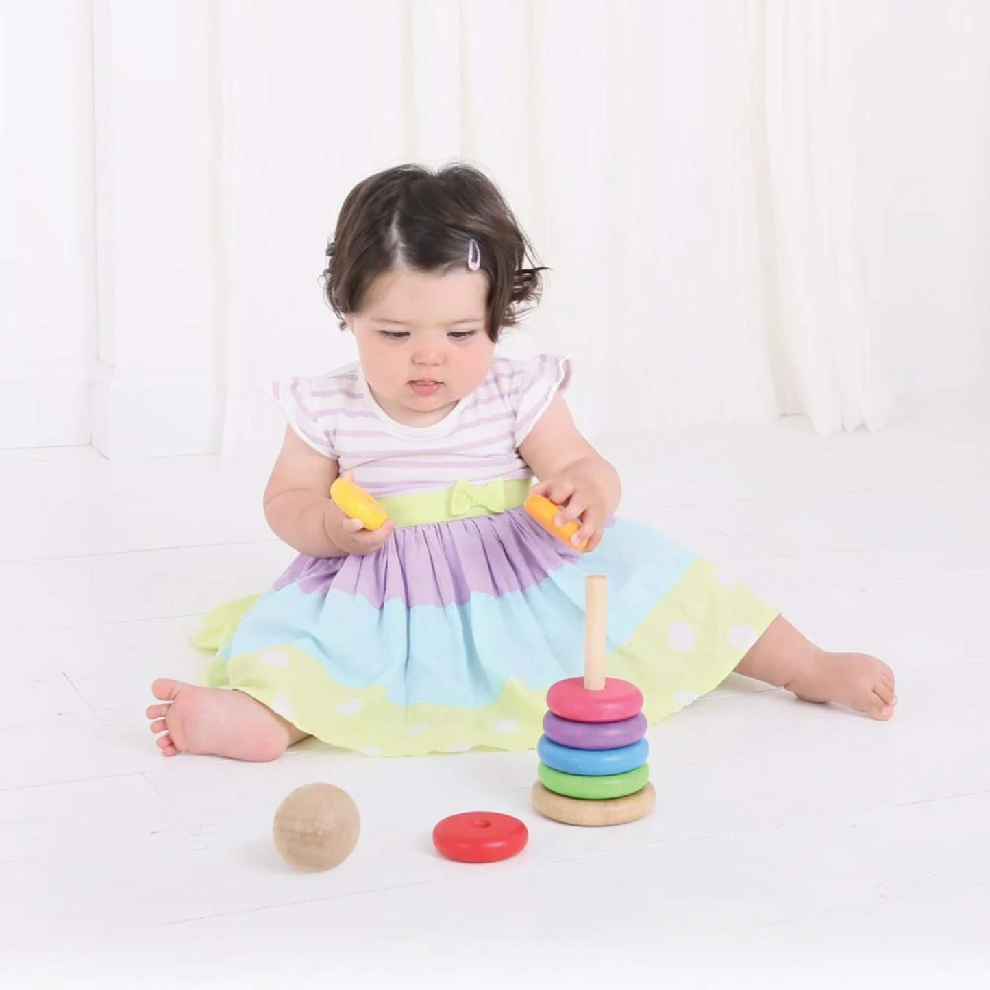 Baby in a pastel dress exploring the wooden rainbow stacker, holding two yellow rings.