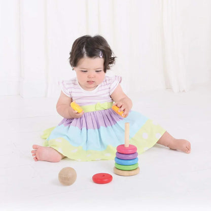 Baby in a pastel dress exploring the wooden rainbow stacker, holding two yellow rings.