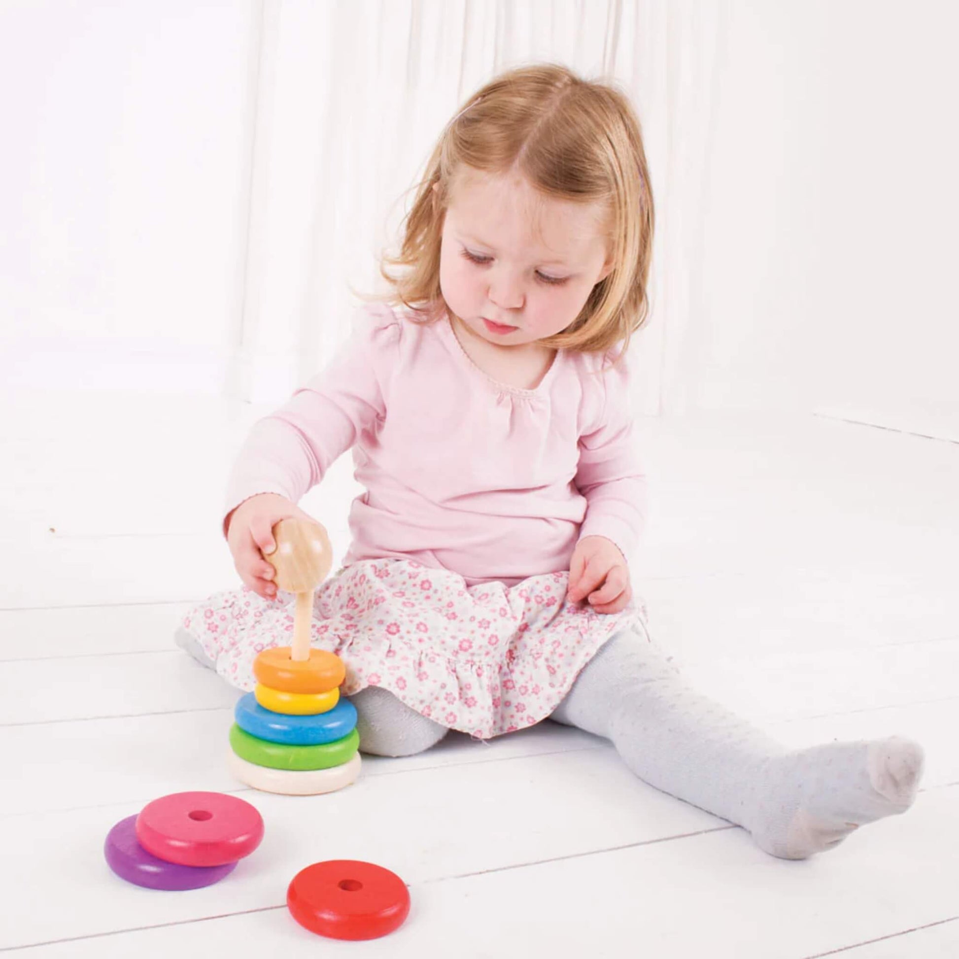 Toddler in a pink top and floral skirt carefully adding a ring to the stacker, with a few pieces nearby.