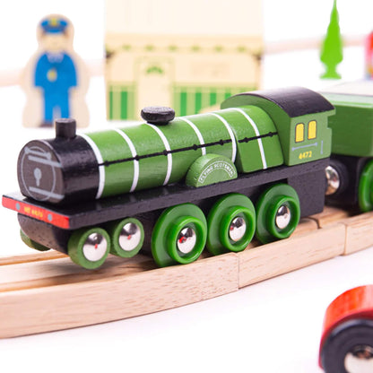 Green Flying Scotsman engine showing white detailing and wheels, positioned on a curved wooden track with station behind.