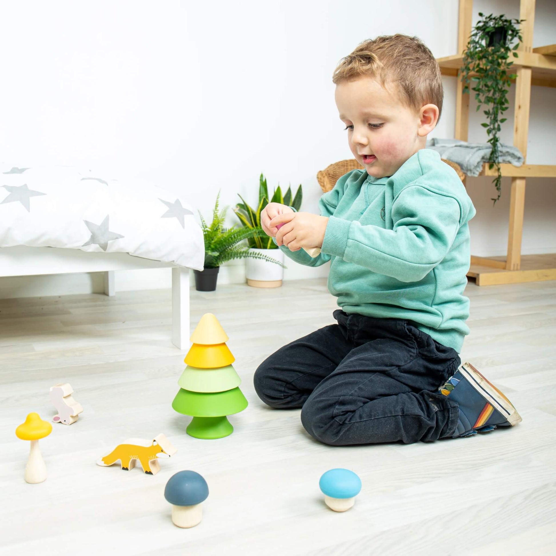 A young child playing on the floor with the Forest Friends Playset, arranging the tree, mushrooms, and animal figures.