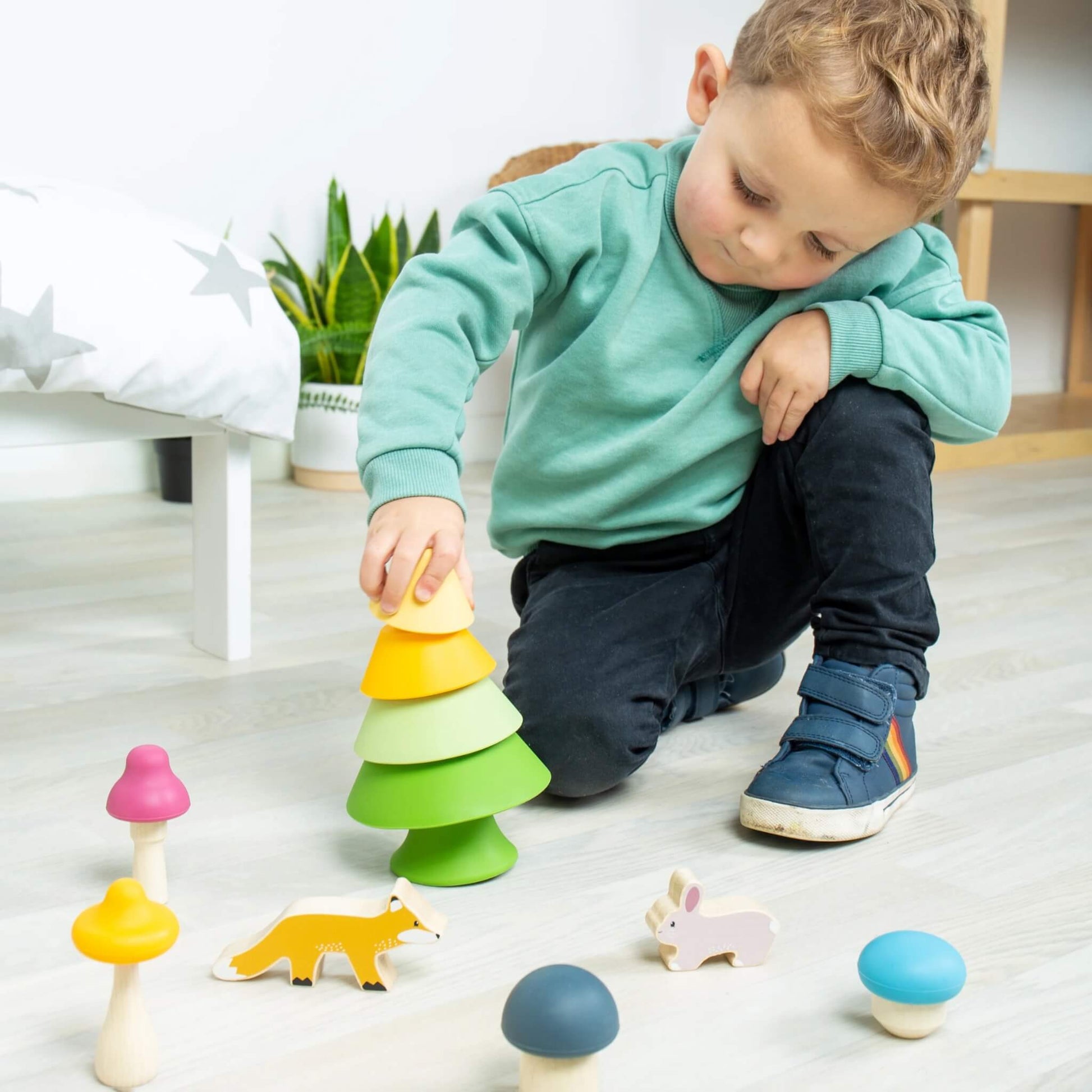 A young child stacking the tree components from the Forest Friends Playset, with the bunny, fox, and mushrooms nearby.