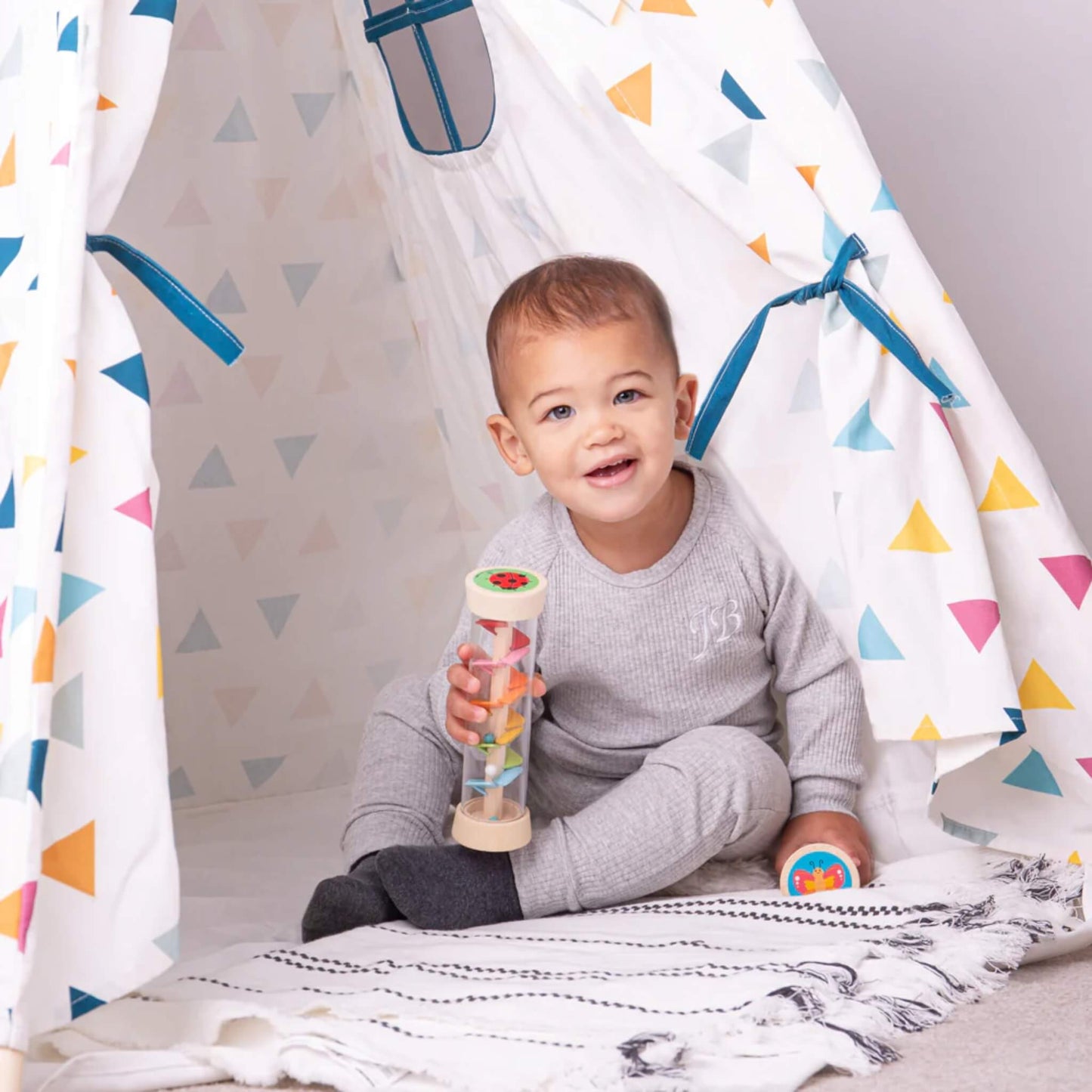 A smiling toddler sits by a play tent holding a wooden Rainmaker, engaging in sensory play on a soft rug.