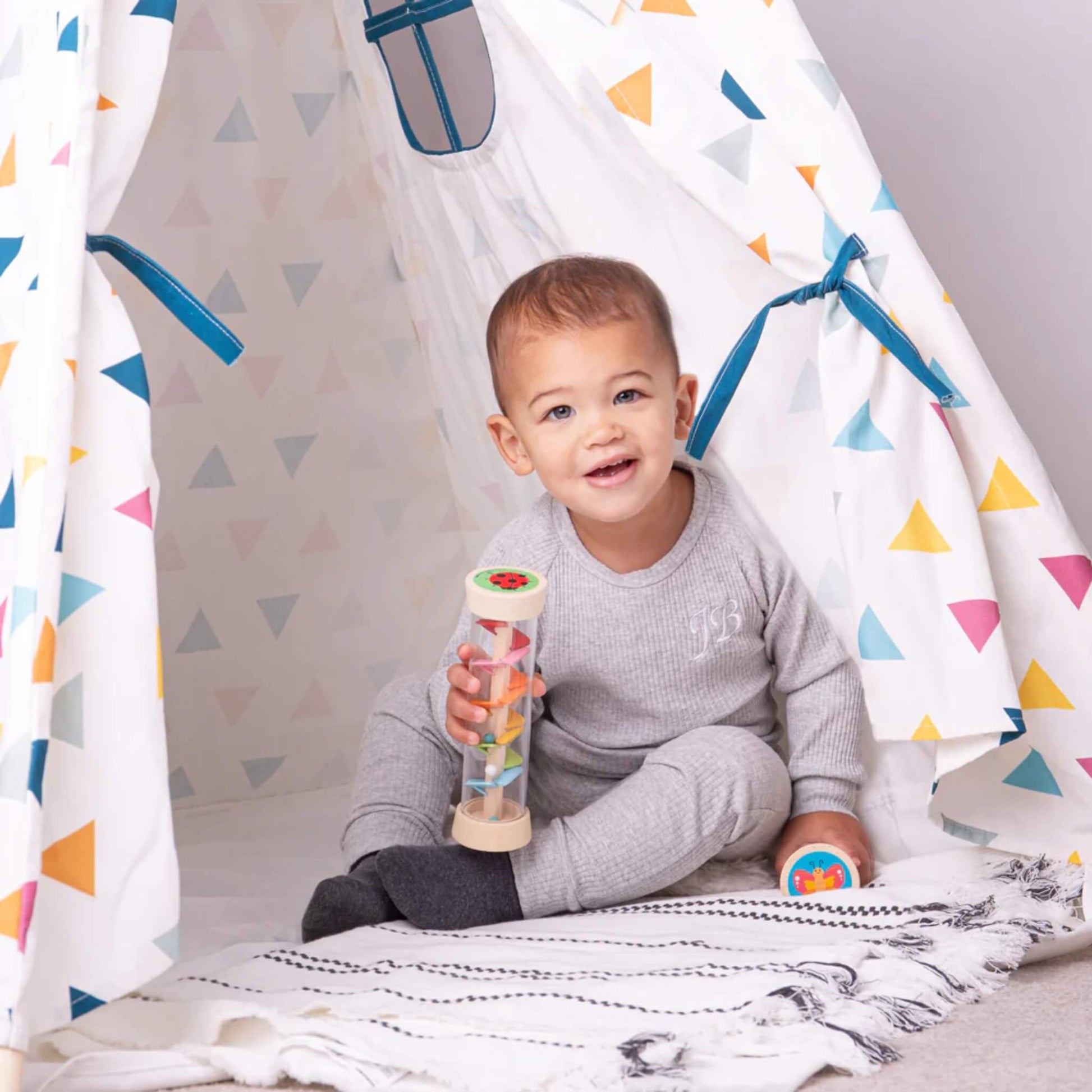 A smiling toddler sits by a play tent holding a wooden Rainmaker, engaging in sensory play on a soft rug.