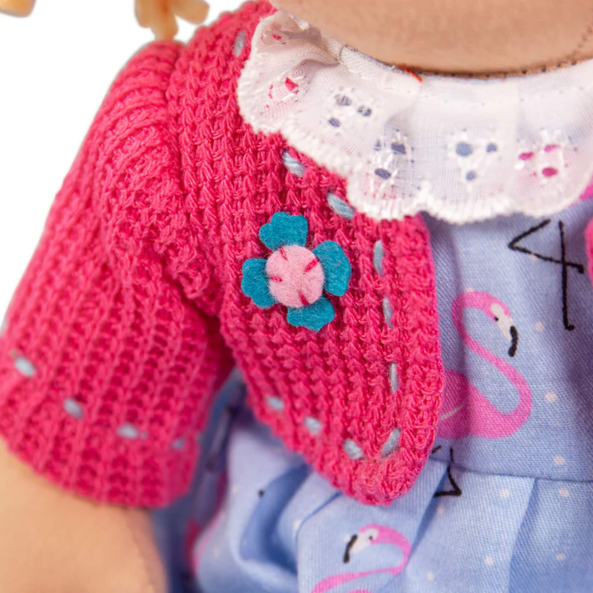 Close-up of the Maggie Doll’s pink knitted cardigan, showing embroidered blue flower embellishment and neat stitched trim.