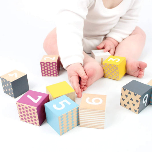 Baby sitting on a white surface, reaching for Bigjigs number blocks scattered around, showing numbers, patterns, and bright colours on wooden cubes.