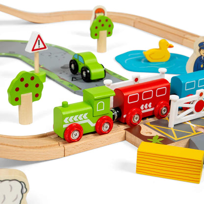 Wooden green train engine and red carriage crossing the level crossing, alongside toy trees, traffic signs, and road pieces from the Bigjigs Road and Rail Train Set.