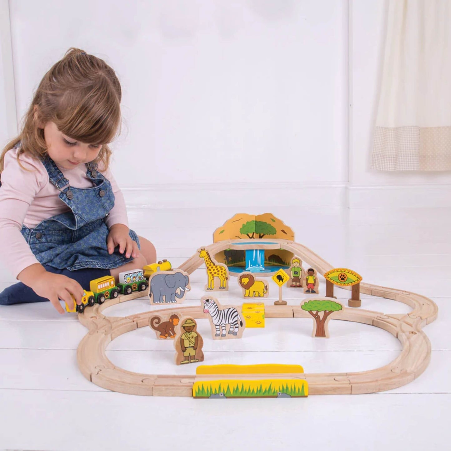 Young girl in a pink top and denim pinafore sitting and playing with the Bigjigs Safari Train Set on a white floor.