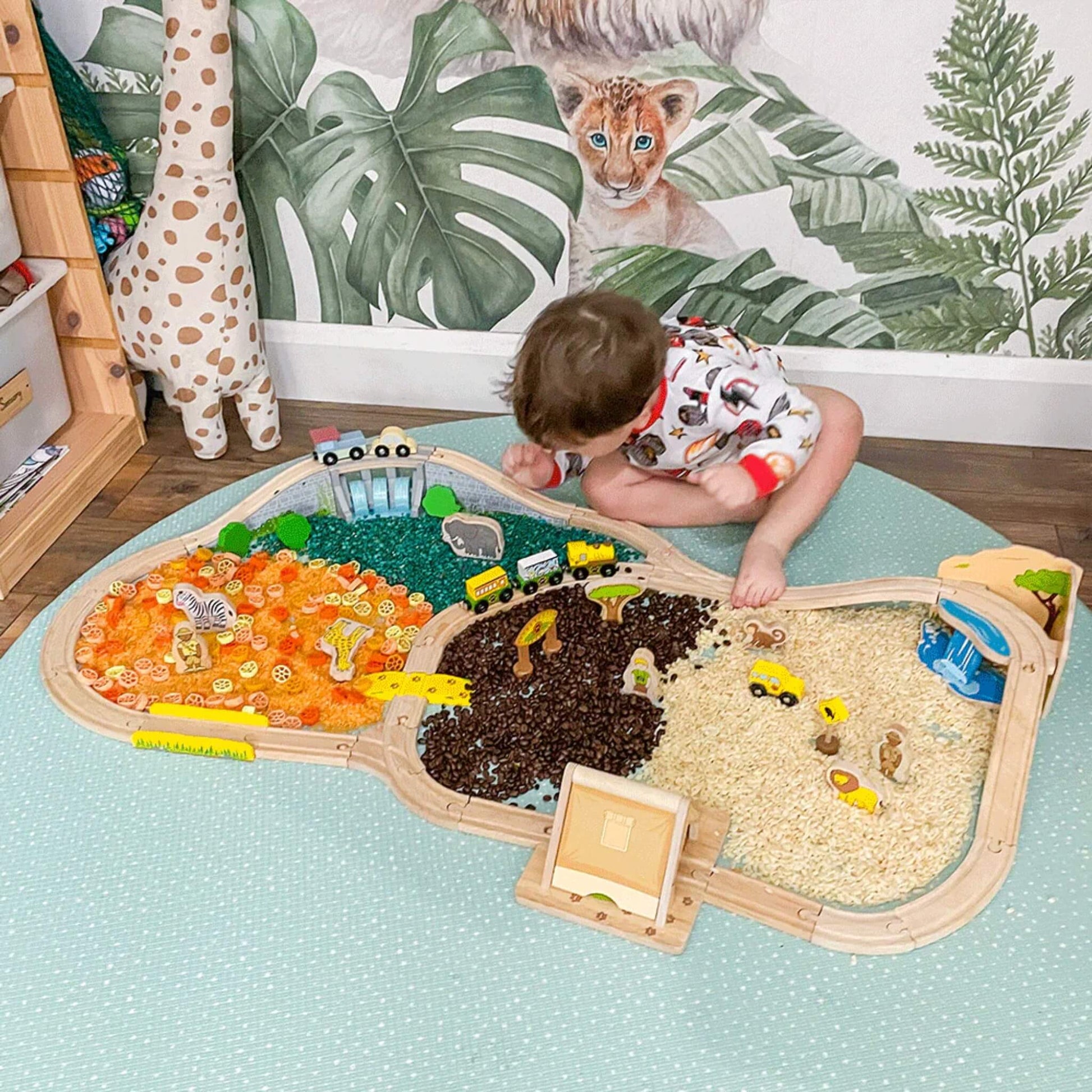 A toddler in printed pyjamas playing with the Safari Train Set on a sensory play mat divided into textured zones for different safari terrains.