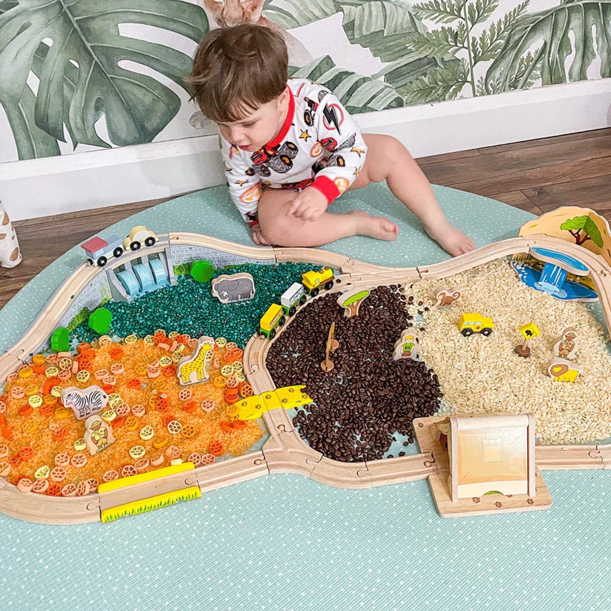 Child sitting by the Safari Train Set displayed on a textured mat, showing animals, safari vehicles, and characters across coloured rice and pasta play areas.
