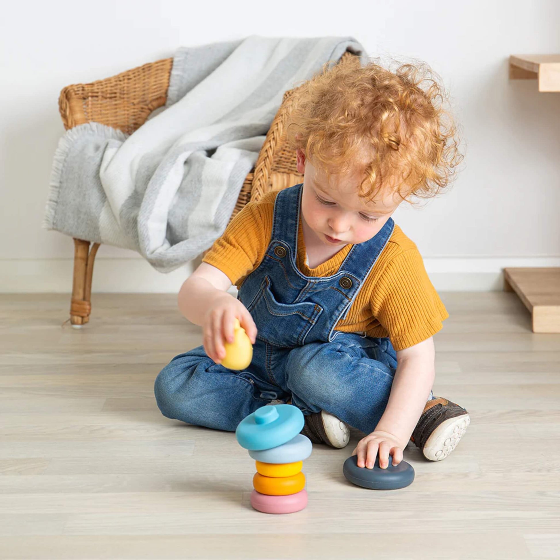 Toddler sitting on the floor in denim overalls, stacking colourful silicone rings from the Bigjigs Toys cat toy, focusing intently on the next piece.