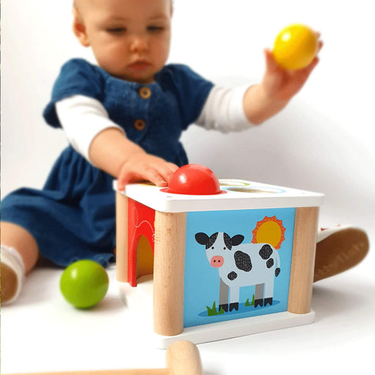 Baby in a blue dress playing with the Bigjigs Toys Tap Tap Ball set, holding a yellow ball and reaching for the red ball on top of the wooden box.