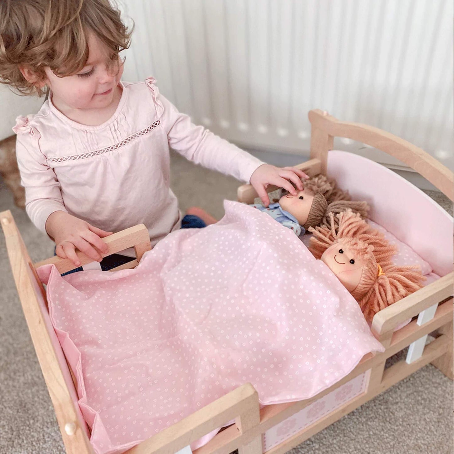 Toddler tucking Zoe Doll and a second ragdoll into a wooden toy bed with pink bedding.