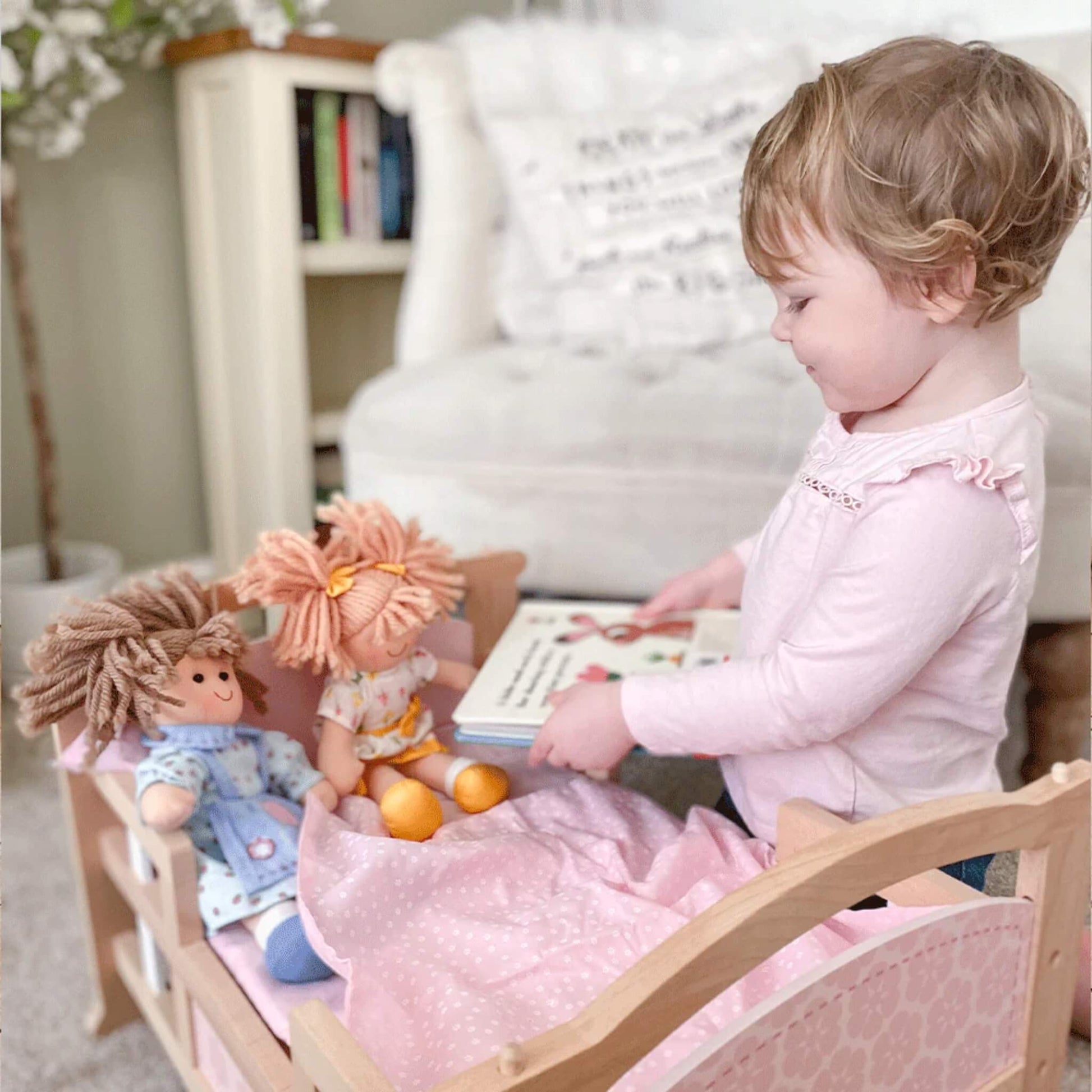 Young girl reading to Zoe Doll and another ragdoll sitting upright in a wooden doll's bed.