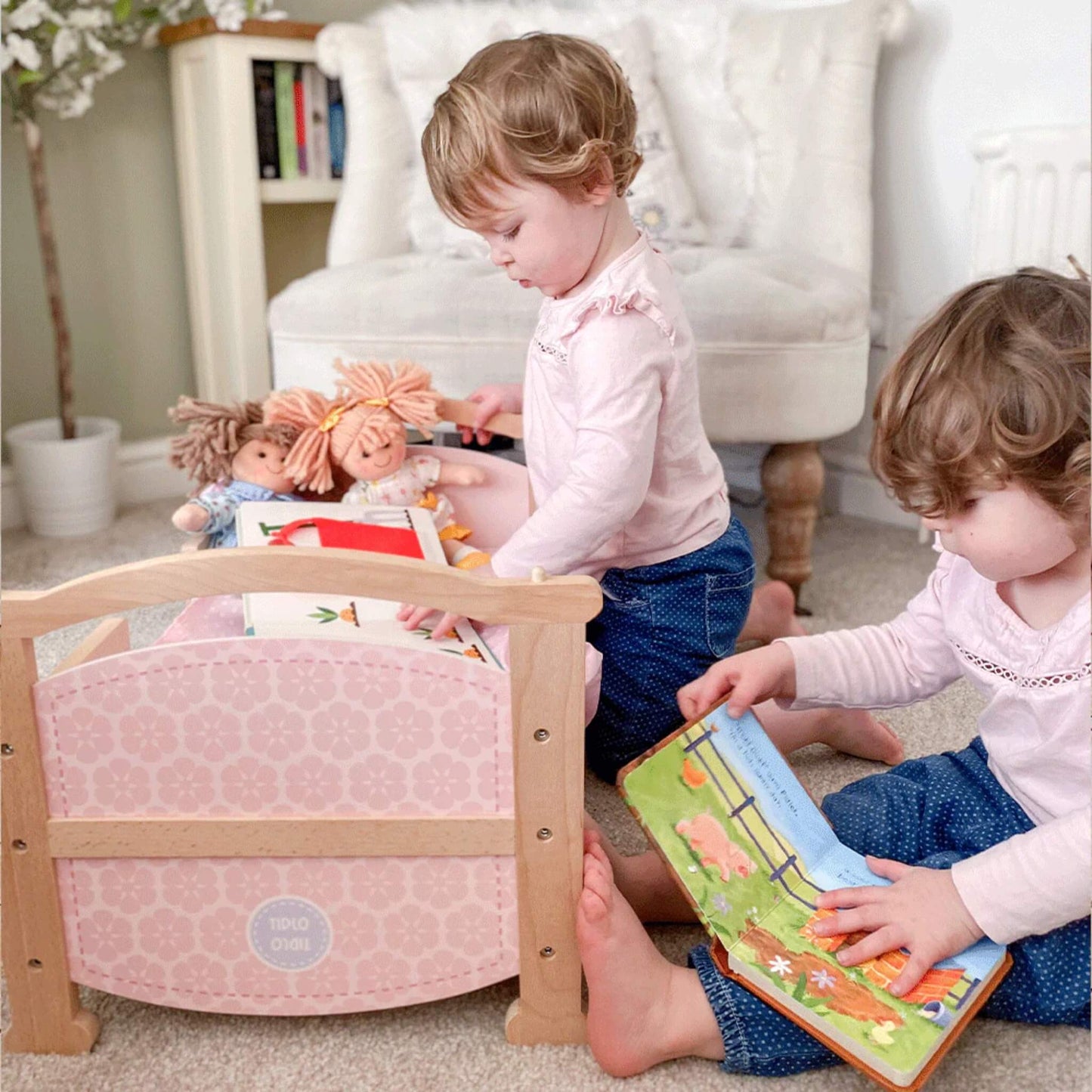 Two toddlers reading books while Zoe Doll and a second ragdoll rest in a wooden toy bed with pale pink quilt.
