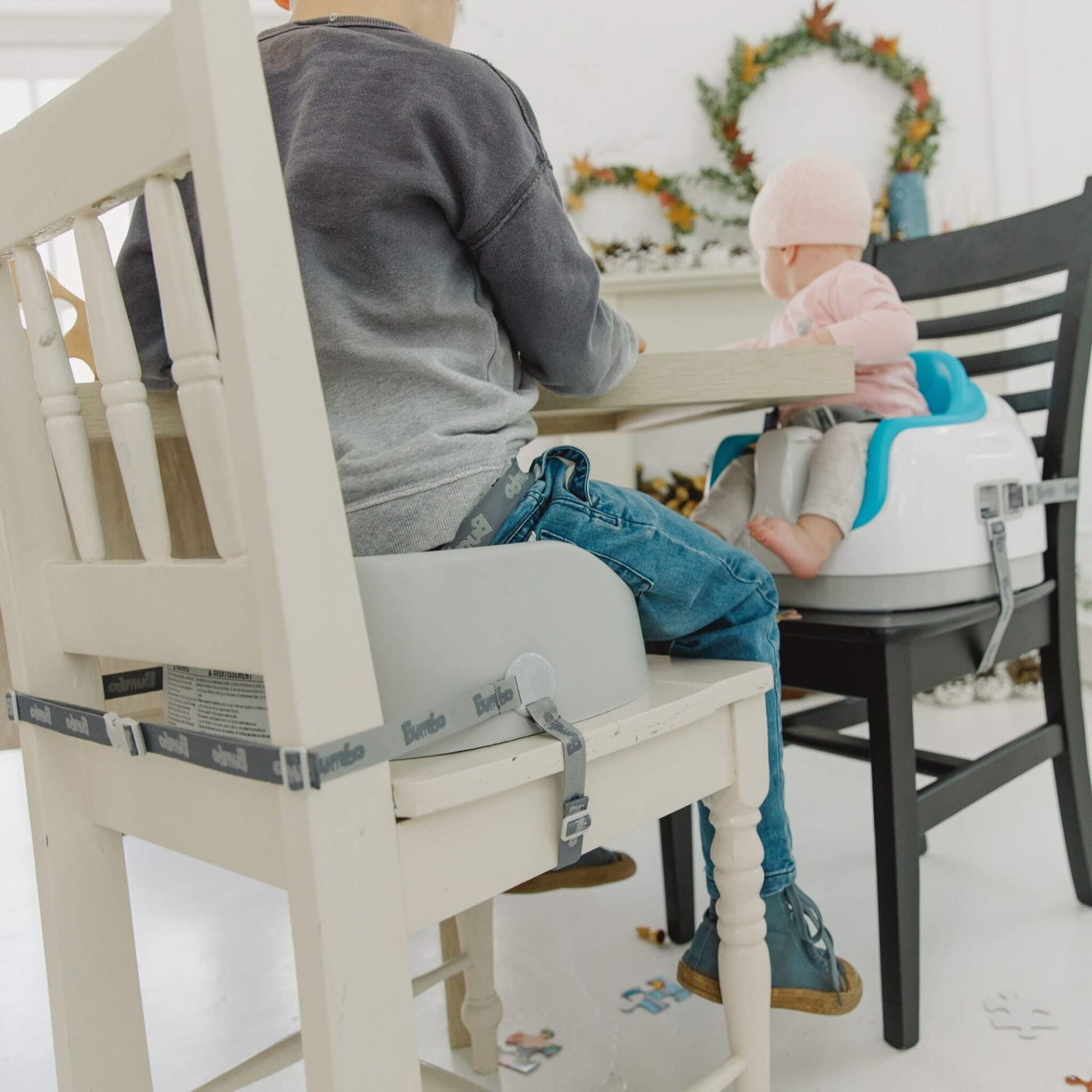 Side-by-side view of two children sitting at a table, one using the Bumbo Booster Seat in Cool Grey on a white chair.