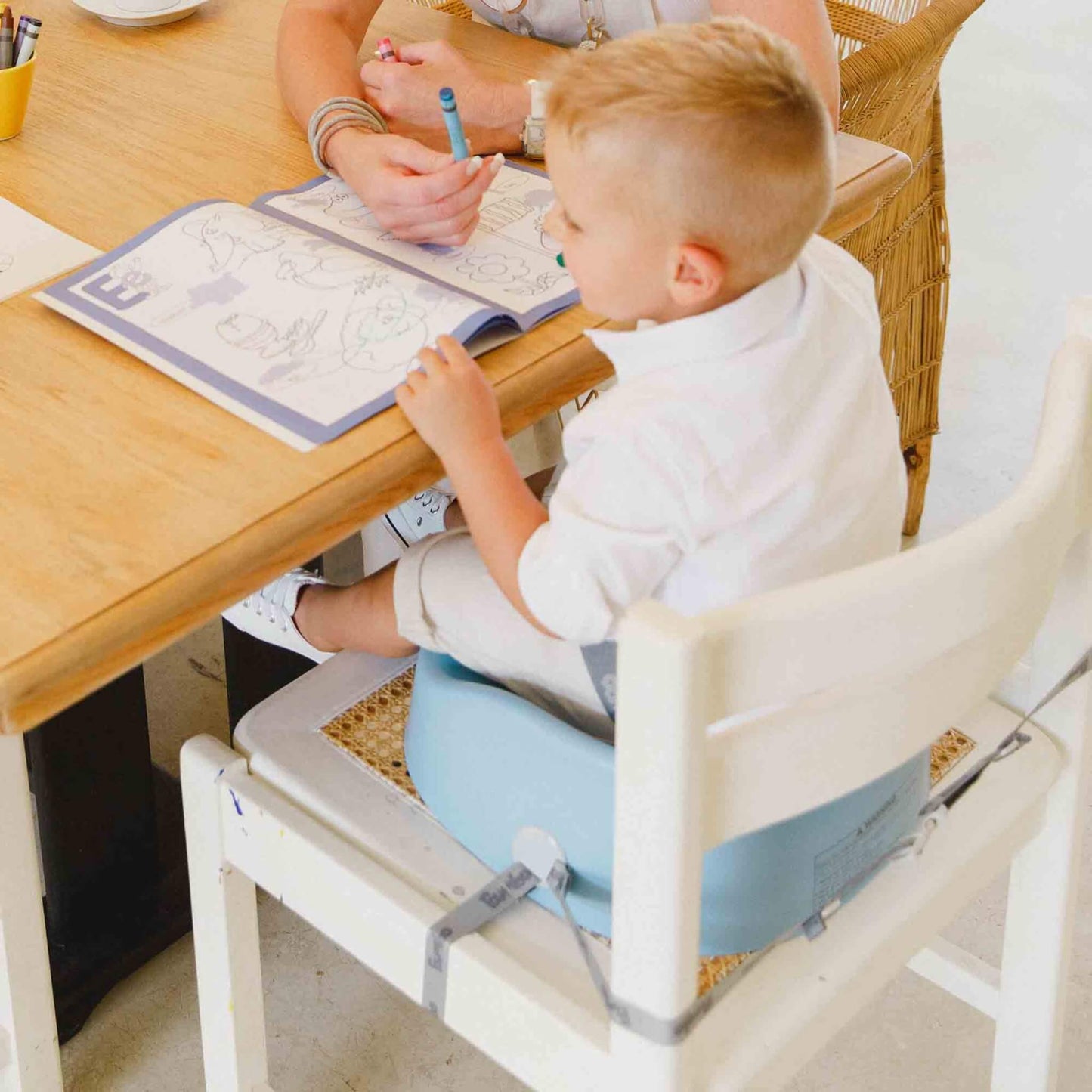 Toddler sitting in the Bumbo Booster Seat in Powder Blue at a wooden table, colouring with an adult, securely fastened to a white dining chair.