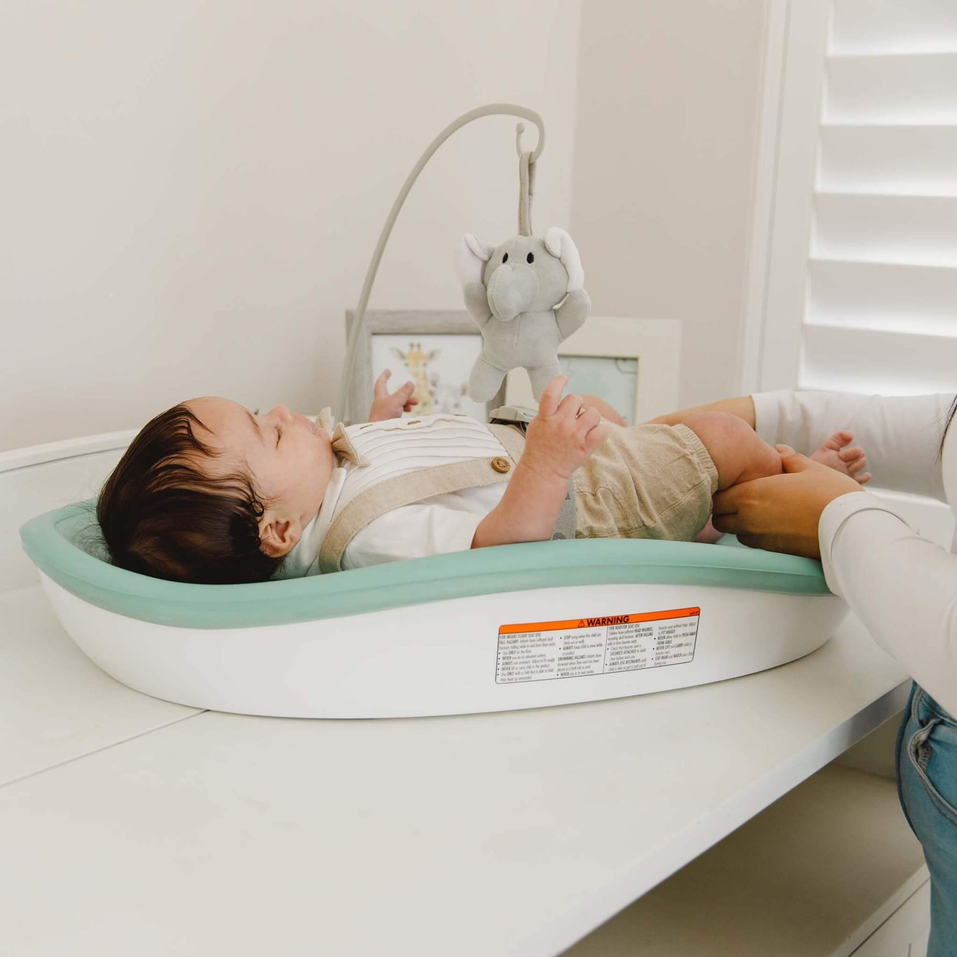 Baby lying on the Bumbo Max Changing Pad in Hemlock, secured with harness and reaching for hanging toy.