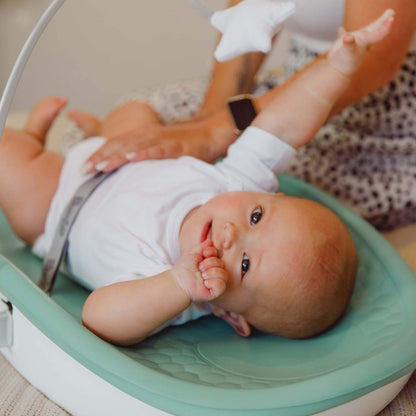 Close-up of smiling baby lying comfortably on the Bumbo Max Changing Pad in Hemlock, secured with safety harness.