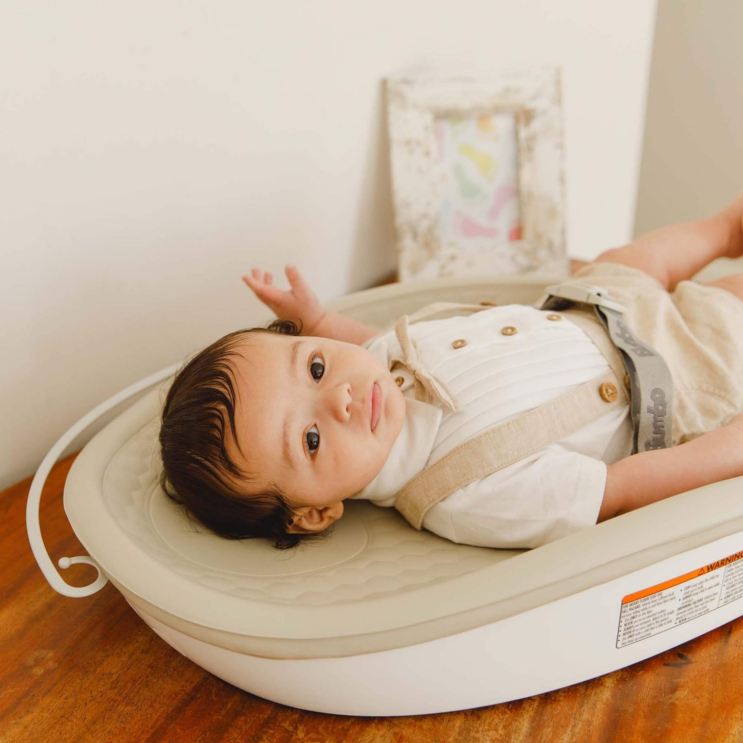 Baby lying on the Bumbo Max Changing Pad in Taupe, secured with harness, placed on a wooden changing table.