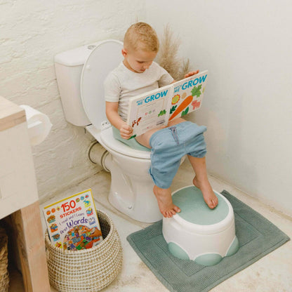 Young child sitting on a toilet using the Step 'n Potty as a footrest, reading a book in a neutral bathroom setting.