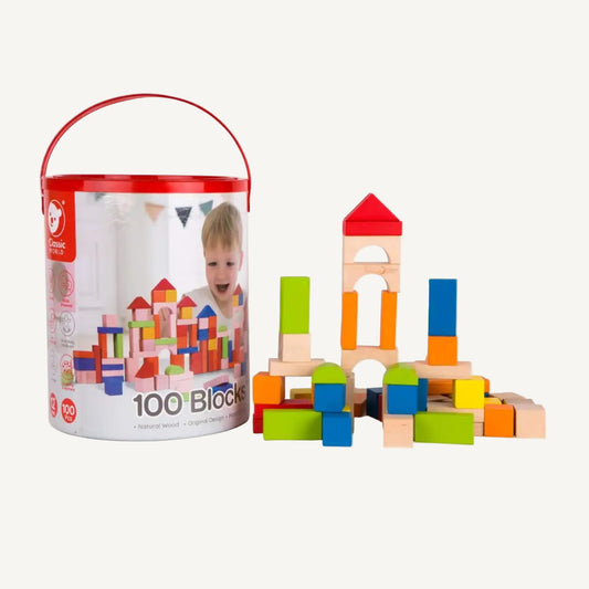 A cylindrical storage tub with red lid and handle featuring printed artwork of wooden blocks, placed beside a small built structure and loose pieces.