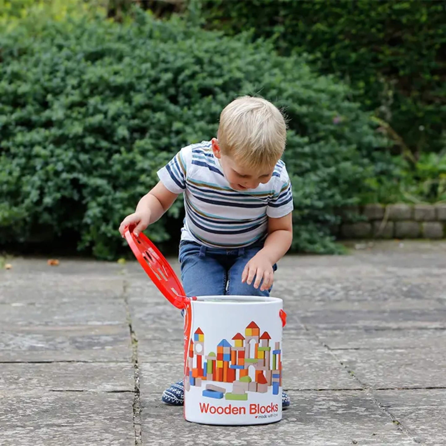 A young child opening a round storage tub of wooden blocks outdoors, reaching inside while standing on a paved surface with greenery behind.