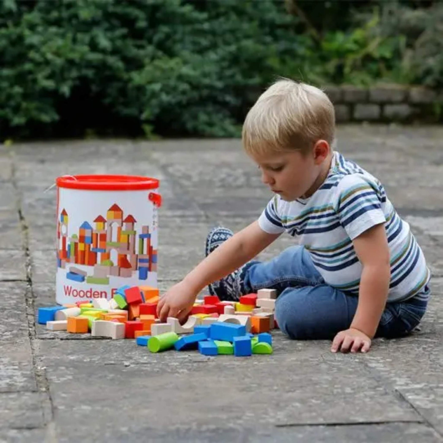 A child sitting on a patio playing with scattered wooden blocks, selecting pieces from a colourful mix of shapes spread across the ground.