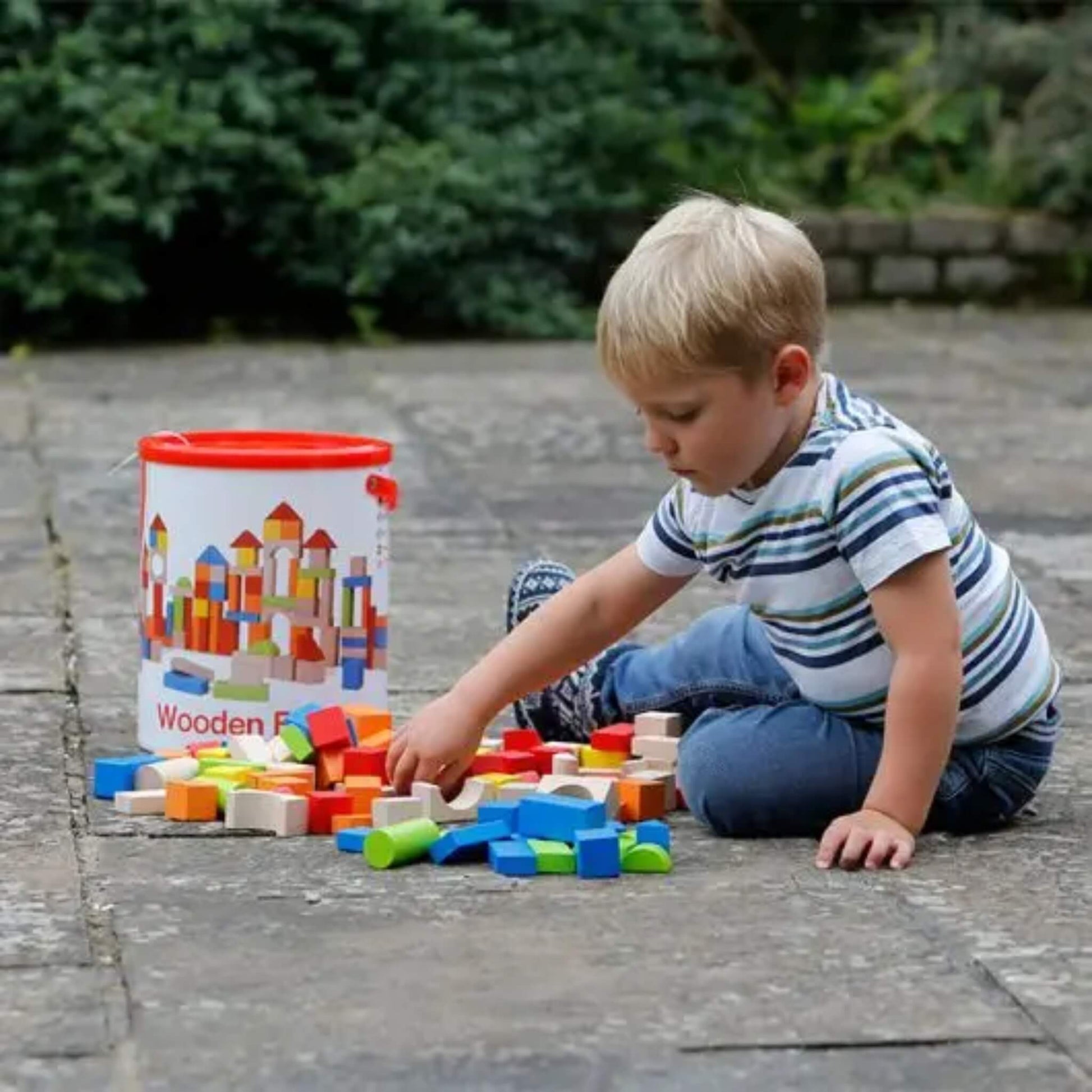 A child sitting on a patio playing with scattered wooden blocks, selecting pieces from a colourful mix of shapes spread across the ground.