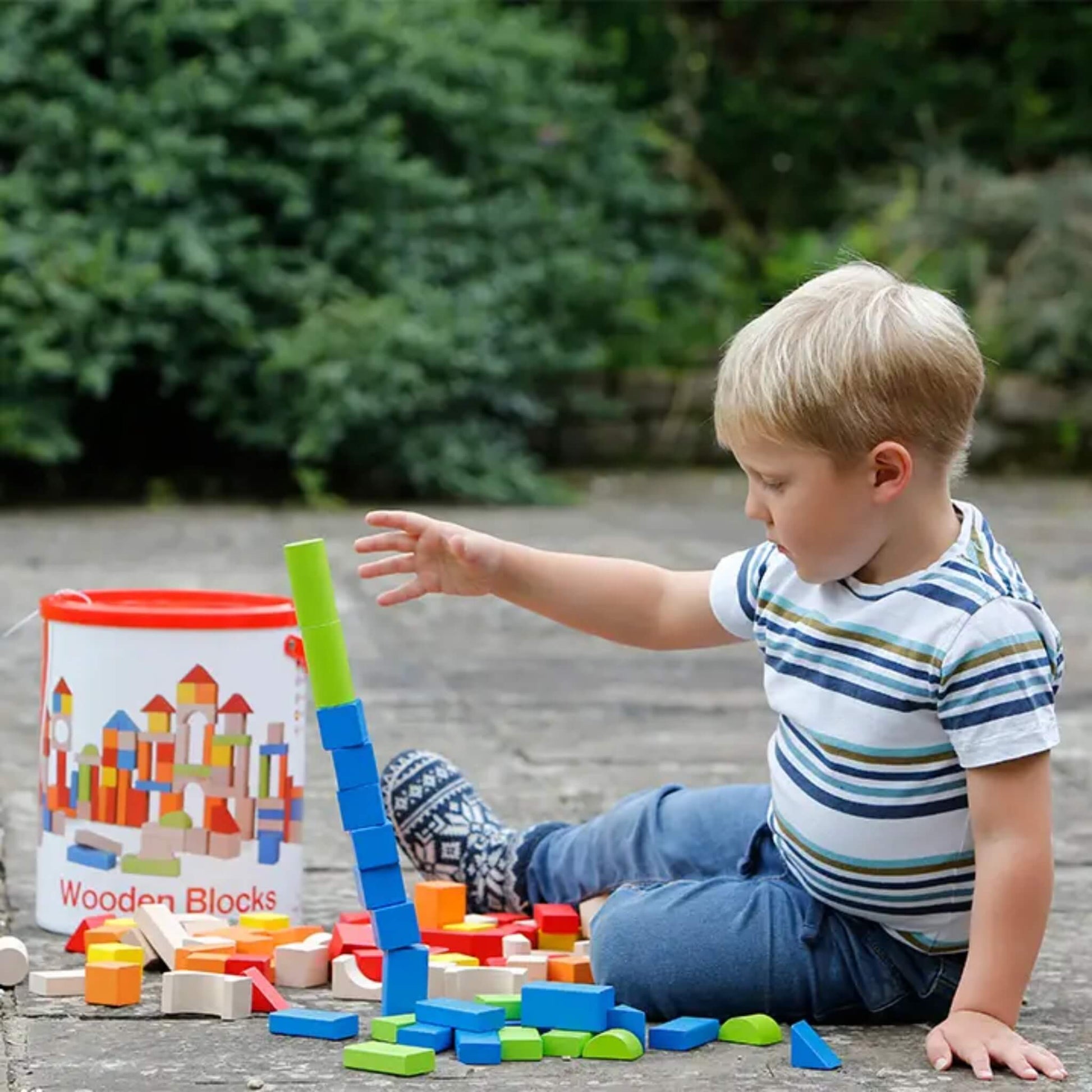 A child stacking tall columns of wooden blocks outdoors, focusing on balancing pieces while sitting beside an open storage tub.