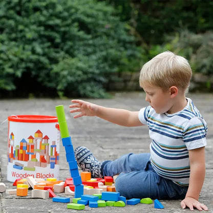 A child stacking tall columns of wooden blocks outdoors, focusing on balancing pieces while sitting beside an open storage tub.