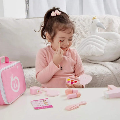 Young girl smiling while pretending to apply wooden toy makeup from a pink beauty set, with play accessories neatly spread out on the table.