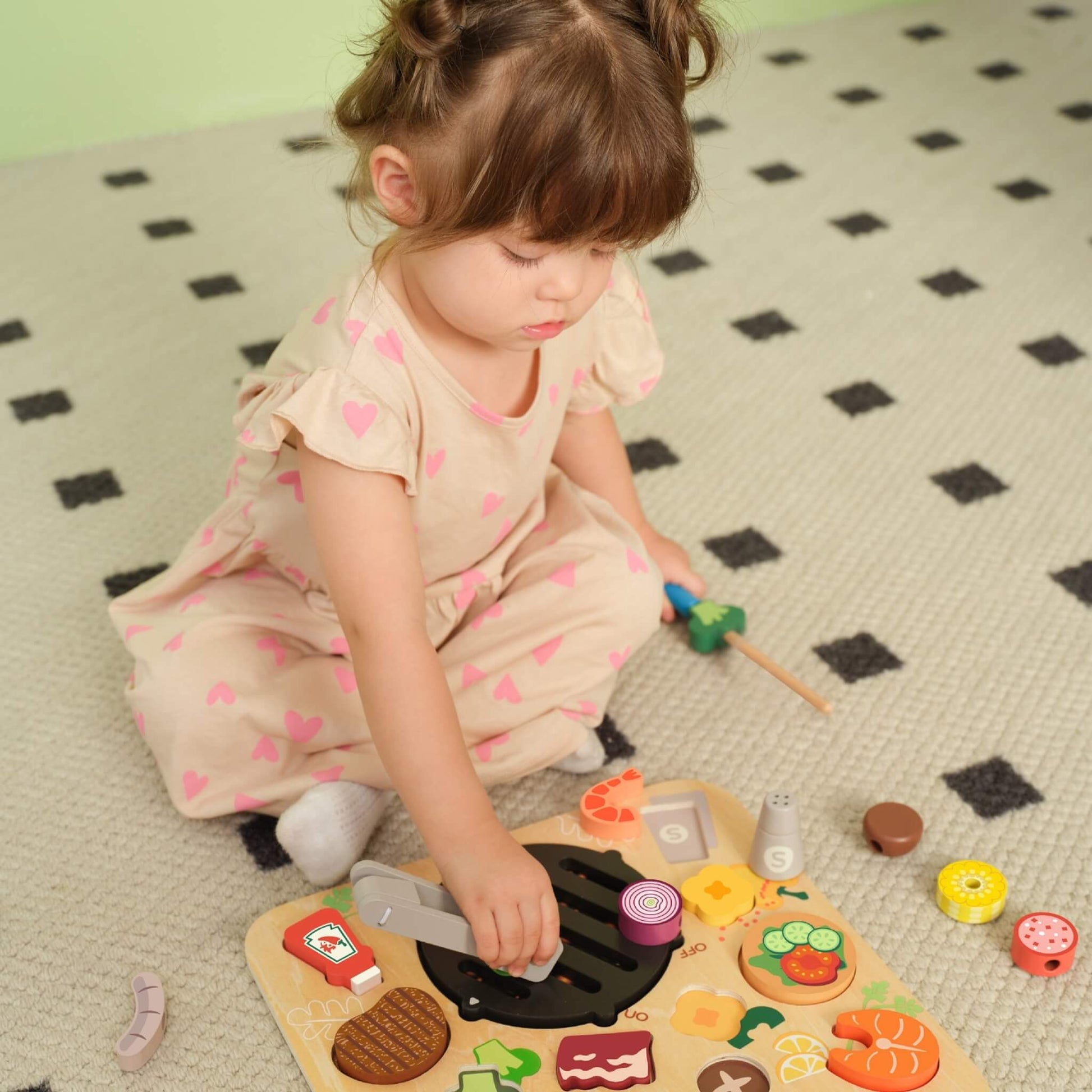 Toddler sitting on a patterned rug using wooden BBQ play set, holding a skewer while placing pieces onto a grill board.