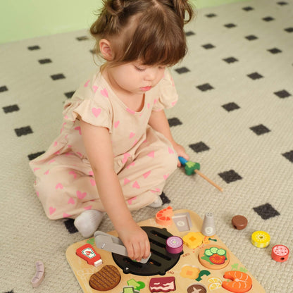 Toddler sitting on a patterned rug using wooden BBQ play set, holding a skewer while placing pieces onto a grill board.